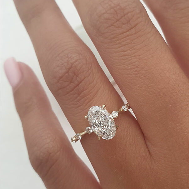 An elongated oval solitaire dainty pave moissanite engagement ring with a gold band.